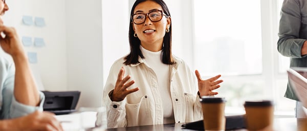 A professional woman wearing glasses and a light-colored outfit speaks during a meeting in a bright office. She gestures with both hands while colleagues listen nearby. Coffee cups and notebooks on the table create a collaborative atmosphere focused on teamwork and discussion.